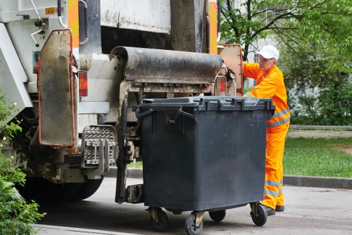 A worker dressed in bright orange high-visibility clothing and a white cap is seen standing next to a large black wheeled rubbish bin, which is positioned in a paved outdoor area. The worker appears to be operating or inspecting the bin, which is situated directly in front of a side loading waste collection truck. The truck has a metal rear section with a partially visible large rolling drum mechanism used for lifting and emptying bins, and the vehicle's housing features areas of exposed metal, weathered surfaces, and safety reflectors. In the background, green trees and a grassy verge are visible, suggesting the scene takes place in a residential or suburban environment of Surbiton or nearby, within the postcode area. The overall scene depicts the process of waste collection in a typical UK town, with a focus on rubbish removal services provided by Commercial Waste Removal Surbiton, emphasizing professional municipal or commercial waste disposal activities.