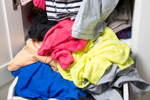 A partially opened white storage drawer filled with a disorganized pile of various clothing items, including brightly coloured T-shirts, a yellow hoodie, grey garments, and a blue piece of clothing, with the clothes appearing crumpled and unused. The drawer is situated on a light-colored surface, possibly within a bedroom or closet, with other similar drawers faintly visible nearby. The setting suggests a domestic environment, typical of a home laundry or wardrobe storage area, which may require waste removal services for decluttering or disposal. The image conveys a casual scene of laundry or wardrobe organisation, with natural lighting illuminating the fabrics' textures and colours, supporting the context of household rubbish or clothing disposal in the Surbiton area.