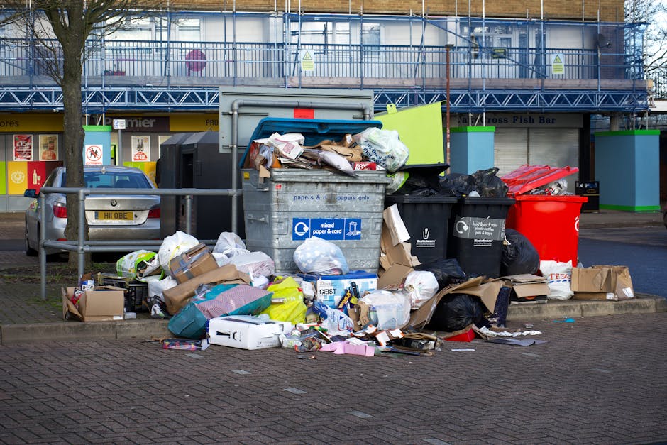 A waste collection worker dressed in a yellow high-visibility vest and dark trousers is positioned at the rear of a large, red rubbish truck, which is parked on the side of a paved urban street. The w