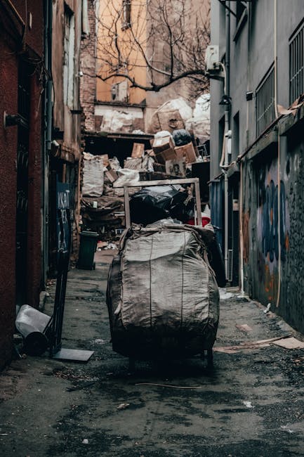 A large collection of mixed household rubbish and packaging waste is piled on a paved sidewalk in front of a parking area, with several black, grey, and red waste bins and a partially overflowing grey