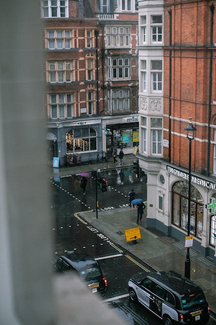 View from an upper-floor window overlooking a rainy city street, with pedestrians holding umbrellas walking across a wet road surface. In the foreground, part of a blurred window frame is visible on t